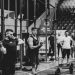 Black and white image of a fitness competition showcasing athletes and weights in a gym setting.